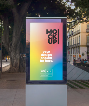Vertical billboard mockup with colorful design, standing on a sidewalk near trees and buildings.
